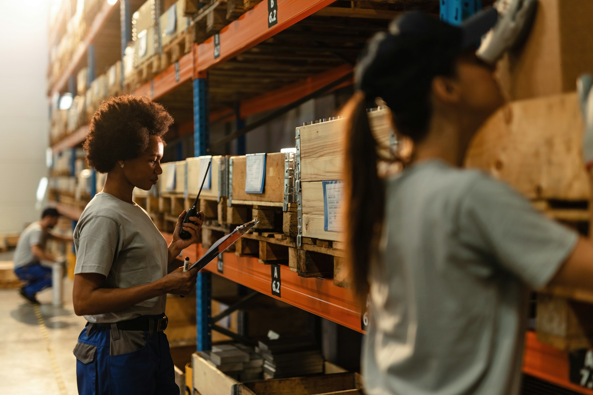 African American warehouse worker communicating on walkie-talkie while going through order list.