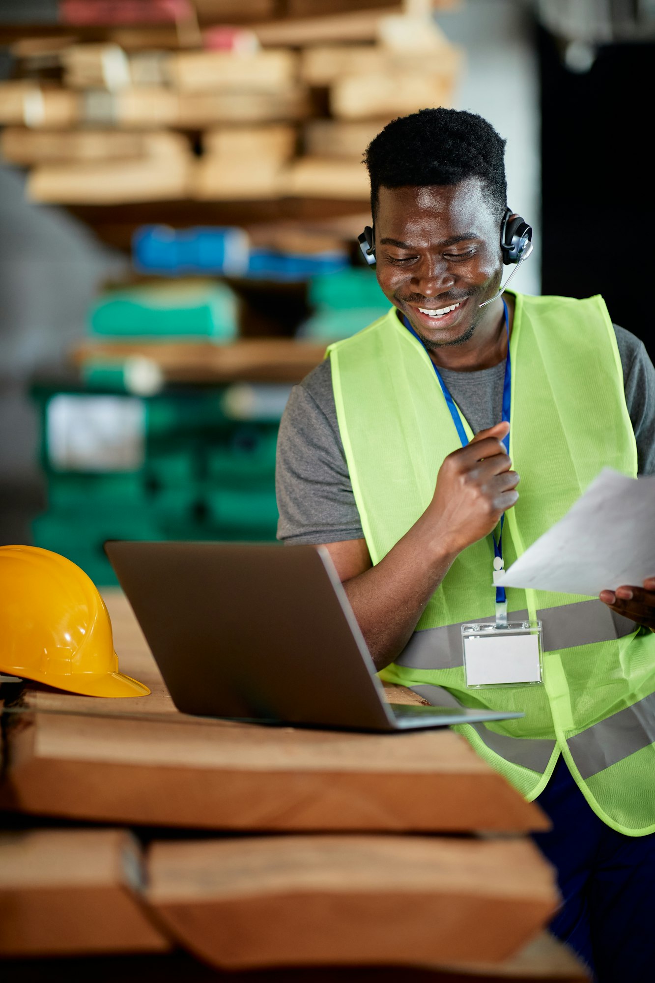 Happy black warehouse worker using laptop and talking via headset while working at wood compartment,